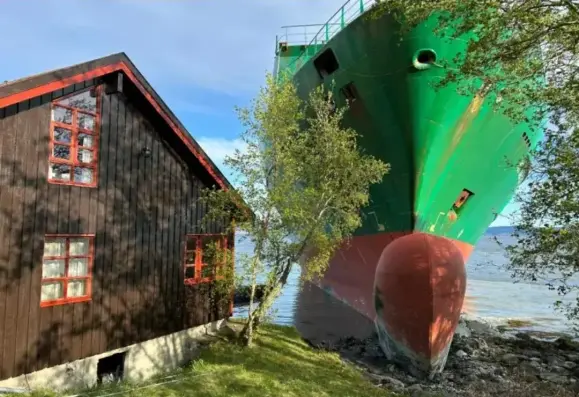Photograph, on the left hand side is a quaint brown cottage with red-orange accents.  On the right, maybe 15 feet away, is the front of a massive green container ship that has run aground and very narrowly missed hitting this house.