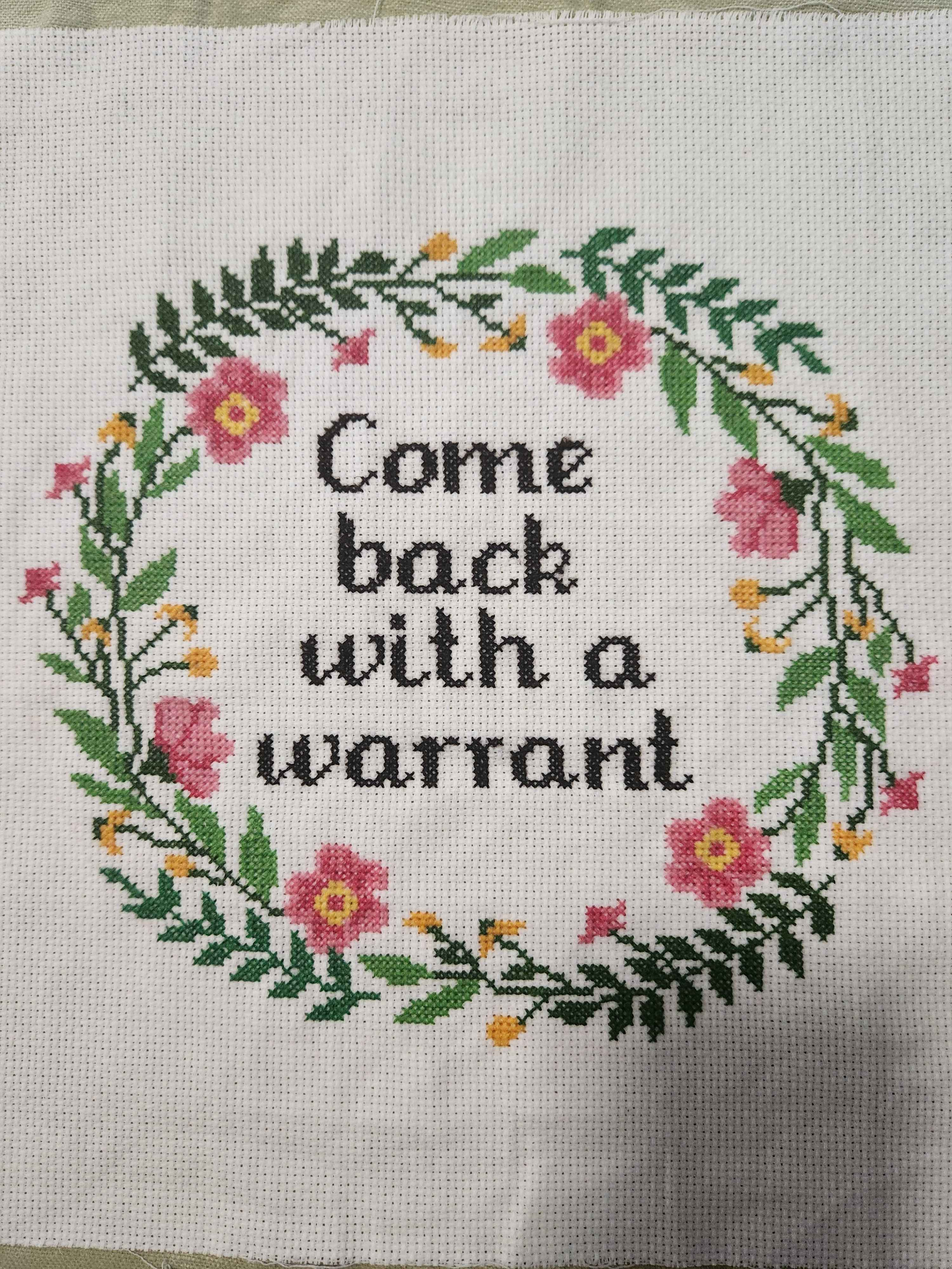 cross-stitch. Black text on a white background: "Come back with a warrant". It is surrounded by a wreath of green leaves and ferns with pink and yellow flowers. It is slightly wrinkly because I just washed it and haven't mounted it yet.