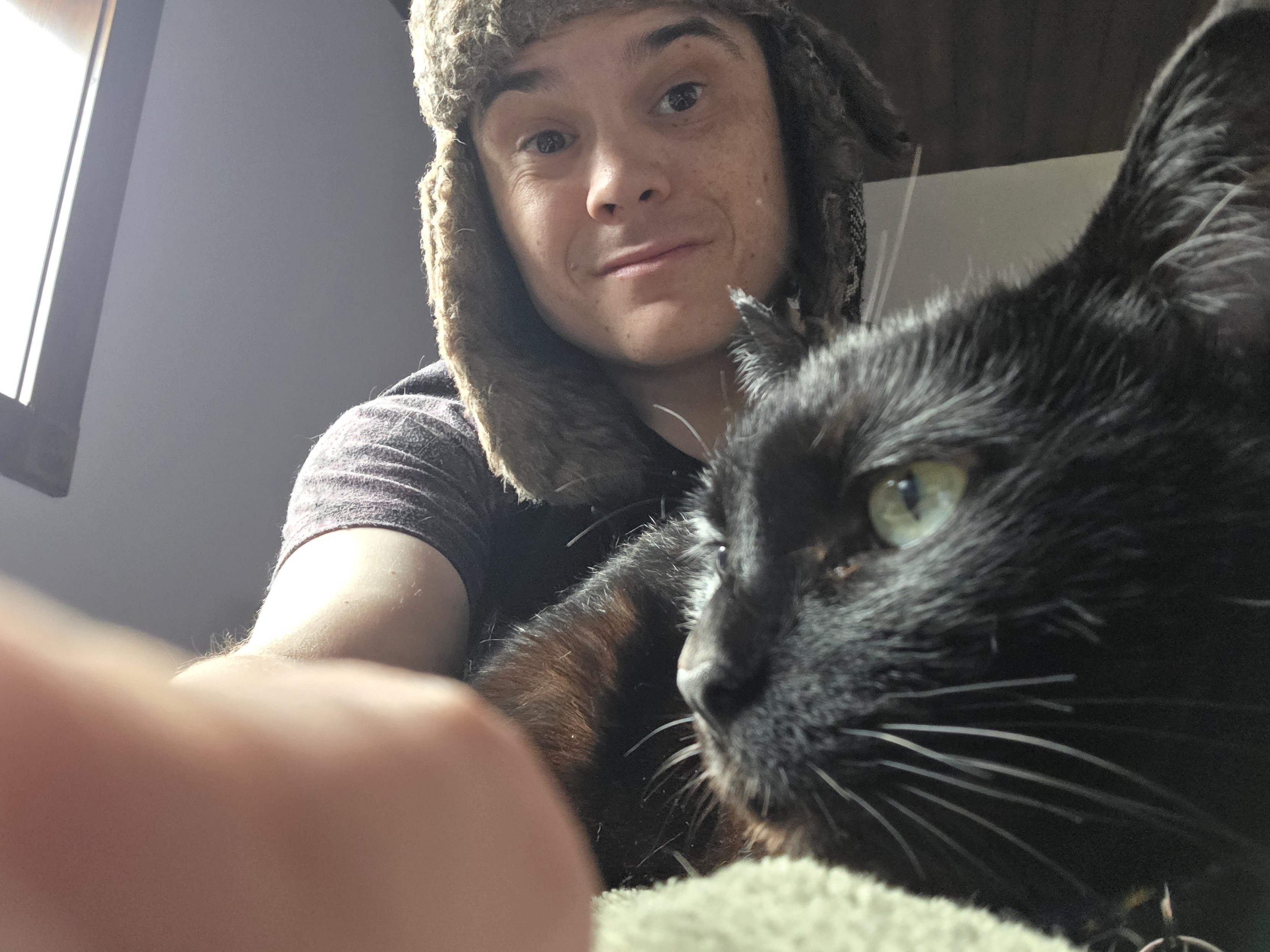 rye smiling, wearing an ushanka and tee, reaching toward the camera. in their lap is her black cat, fennec; you can see here profile